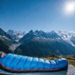Persona durmiendo en un saco de dormir azul haciendo vivac frente a montañas nevadas bajo las estrellas.