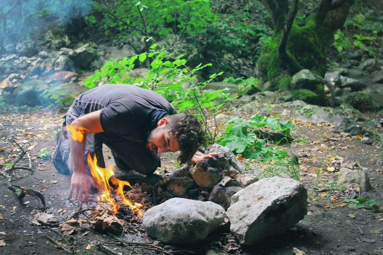 Man attempts to start a campfire surrounded by rocks in a lush green forest.