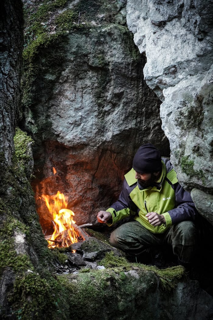 Man in green jacket lights campfire in rocky mountain shelter for survival.
